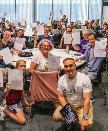 A group of people of all ages in a classroom, smiling and holding paper in the air