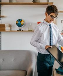 A boy in a school uniform in a home setting, putting a laptop into a backpack