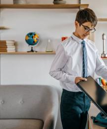 A boy in a school uniform in a home setting, putting a laptop into a backpack
