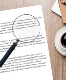 A piece of paper on a desk with text on it. On top of the paper is a magnifying glass, and a cup of coffee is next to it