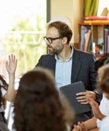 A teacher in a class speaking to a pupil, with the class sitting in a circle. The pupil, a girl, has her hand raised