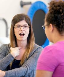 A woman in a therapy setting, pointing at her throat and opening her mouth. Opposite her is a younger woman