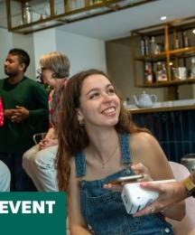 A young man and a woman sitting in a coffee shop, smiling at a woman who is standing beside them with a card payment reader