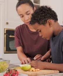 A boy cutting up a lime on a chopping board with his mother looking on. In the background is the father