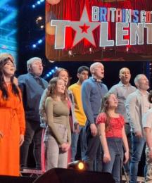 Members of a choir of mixed ages singing on a stage in front of a conductor. Above them is a logo saying 'Britain's Got Talent'