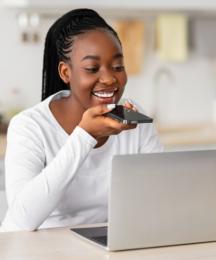 A woman sitting at a kitchen table in front of a laptop and speaking into a smartphone in her hand