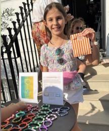 A girl smiling and holding up a paper bag, standing at a stall displaying bracelets