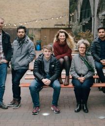 A group of people standing beside and sitting on a bench outdoors and looking at the camera