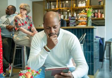 A man in a coffee shop looking at a tablet with his hand to his mouth