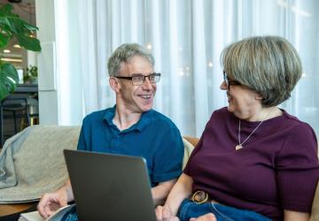 A man and a woman sitting on a sofa laughing, the woman with a laptop on her lap
