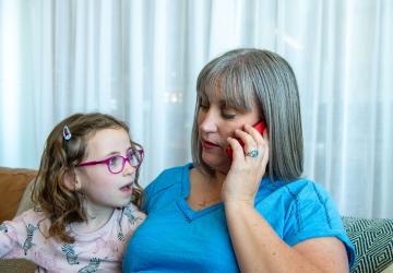 A mother sitting on a sofa and speaking into a phone, with her young daughter sitting next to her