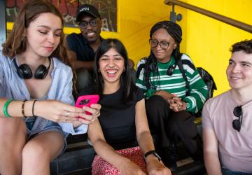 A group of five young people sitting on steps and looking at a phone that one of them is holding, and smiling