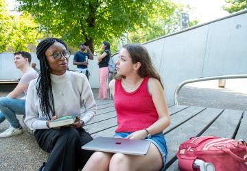 Two women seated in an outside setting and chatting, with other people in the background