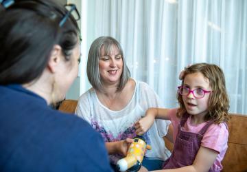 A young girl holding a toy and sitting with her mother while talking to a woman sitting opposite them