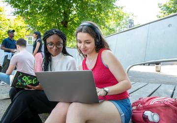 A woman in headphones showing something on her laptop to the woman sitting next to her.