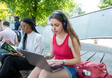 A woman wearing headphones and sitting outside typing on a laptop. Another woman reading a book is sitting next to her.
