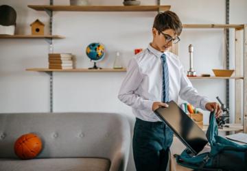 A boy in a school uniform in a home setting, putting a laptop into a backpack