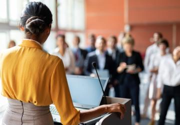 A woman standing a podium and speaking to an audience of people who are standing up