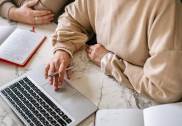 A typing on a laptop whilst holding a pen. Beside her is an other person. We can't see their faces.