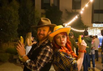 A man and a woman in cowboy outfits standing with their backs to each other and holding up bananas instead of guns