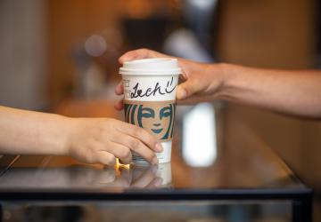 A hand passing a Starbucks cup over to another hand, on the cup is written 'Zach'.