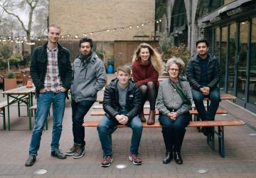 A group of people standing beside and sitting on a bench outdoors and looking at the camera
