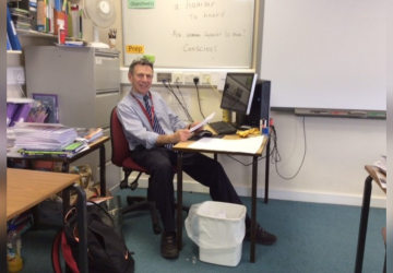 A man sitting at a desk in a classroom, smiling for the camera
