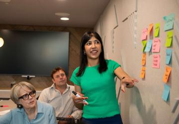 Affiliate With STAMMA A woman in an office standing in front of a series of post it notes on a wall and pointing to them, with two people sitting and watching.