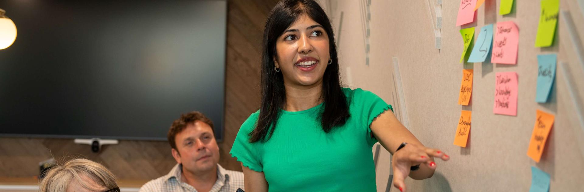 A woman standing next to a wall with post it notes on it, with a woman and a man sitting beside her, watching