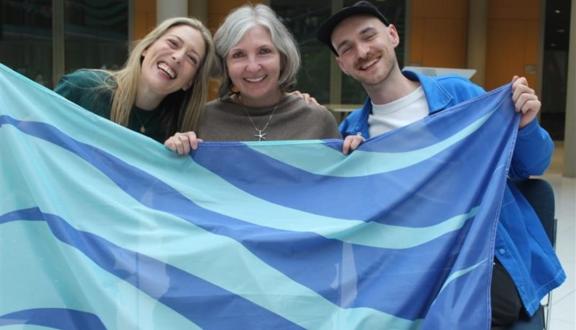Two women and a man smiling and holding a Making Waves stammering pride flag outside