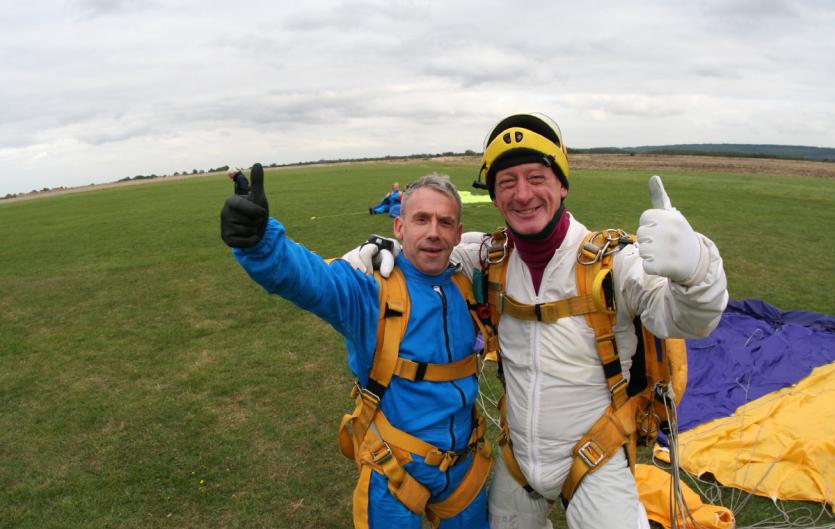 Two men in skydiving outfits looking at the camera with their thumbs up