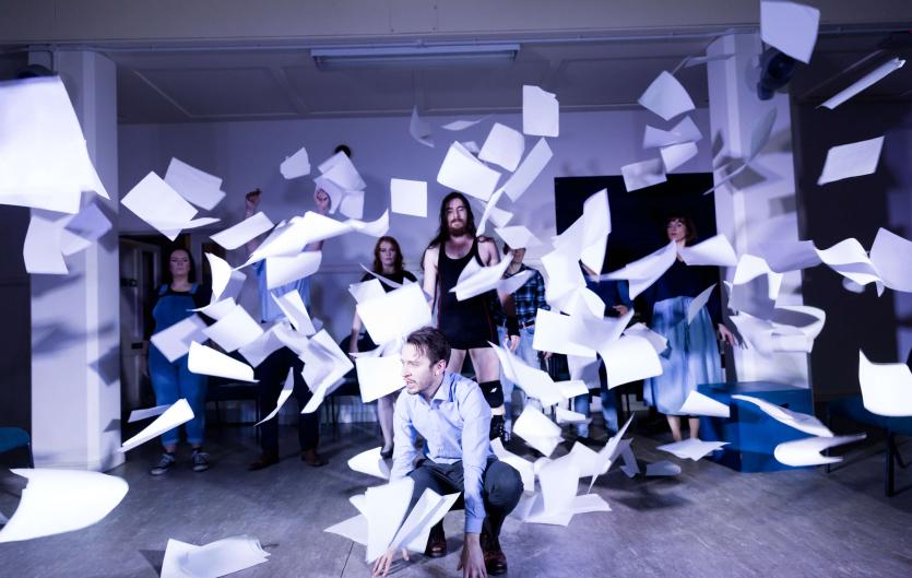 A scene from a theatre play, with a man squatting, a group of people behind him, and paper flying everywhere
