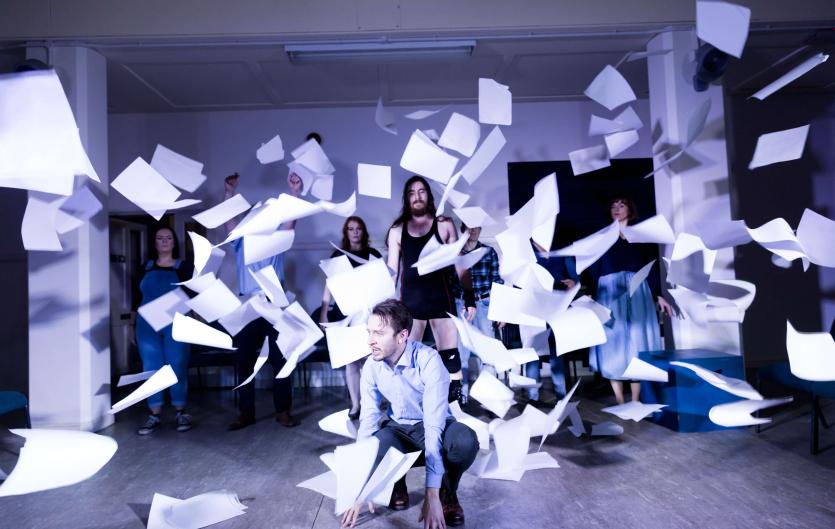 Stammering and the power of theatre A scene from a theatre play, with a man squatting, a group of people behind him, and paper flying everywhere