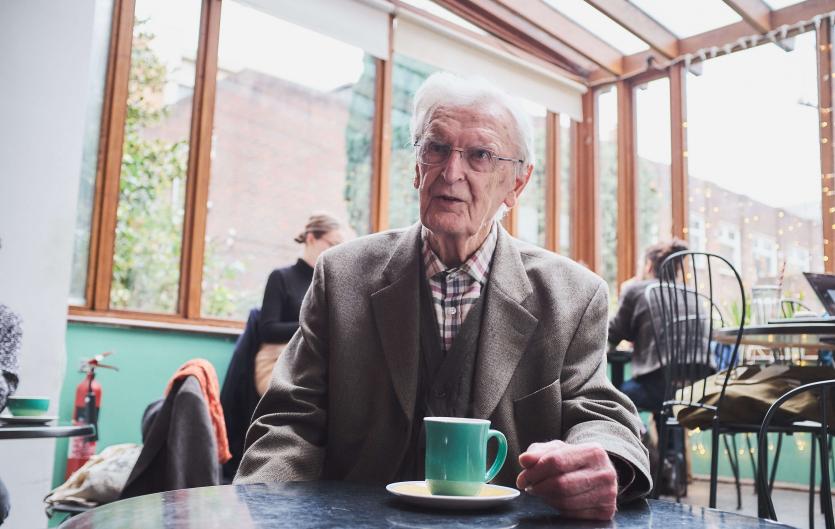 A man in his senior years sitting in a cafe looking to the left.