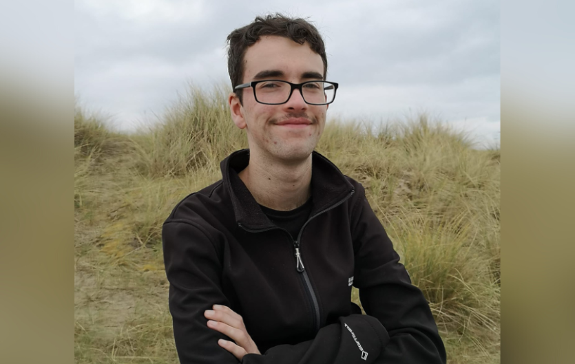 Understanding My Stammer Before University A man looking at the camera and smiling, with his arms folded, with a grassy mound in the background