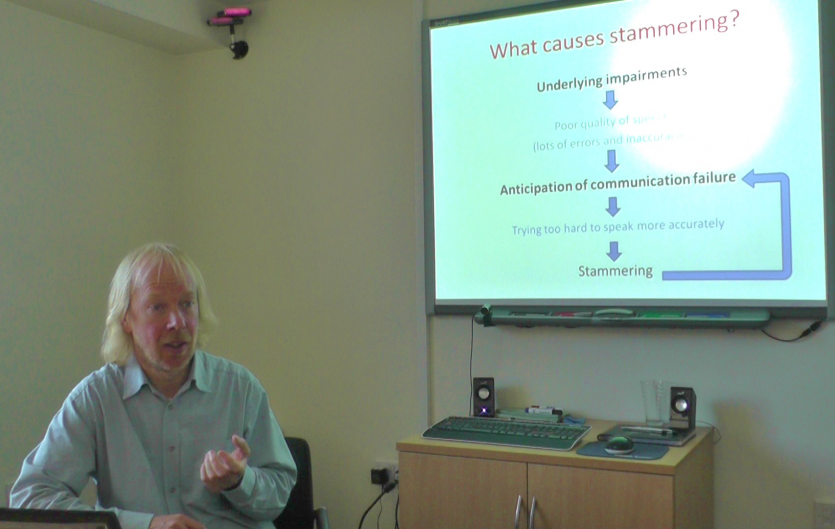 A man giving a talk, with projections on a screen behind him