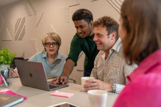 A man standing and demonstrating something on a laptop, watched by a man and a woman sitting either side.