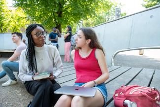 Two women seated in an outside setting and chatting, with other people in the background