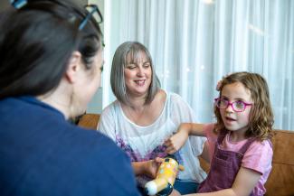 A young girl holding a toy and sitting with her mother while talking to a woman sitting opposite them