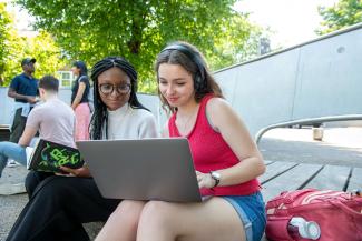 A woman in headphones showing something on her laptop to the woman sitting next to her.