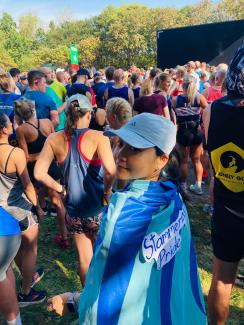 A woman standing in a crowd of people at a charity running event, wearing a cape with a sign on it saying 'Stammering Pride'