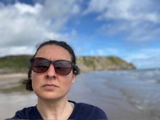 A woman with sunglasses on looking at the camera against a coastal backdrop