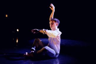A man sitting on a stage, gesticulating with his arms, mid-dance