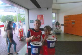 A man and a woman holding collection buckets in the entrance of a supermarket, both looking at the camera and smiling