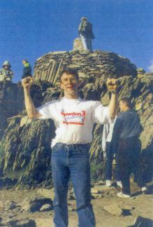 A man on a mountain path holding his arms aloft, looking at the camera and smiling
