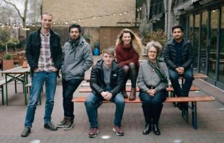 Six people sitting on standing around a bench, looking at the camera