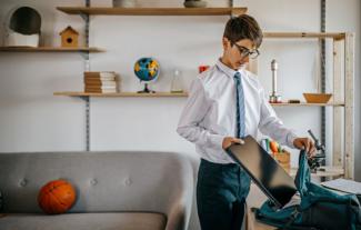 A boy in a school uniform in a home setting, putting a laptop into a backpack