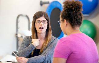 A woman in a therapy setting, pointing at her throat and opening her mouth. Opposite her is a younger woman