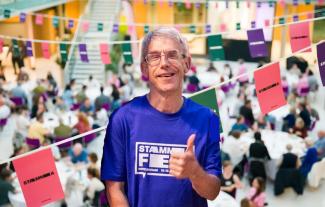 A man smiling and raising his thumb, aganist a backdrop of bunting and a crowded room