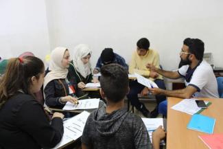 A teacher in a class speaking to a pupil, with the class sitting in a circle. The pupil, a girl, has her hand raised